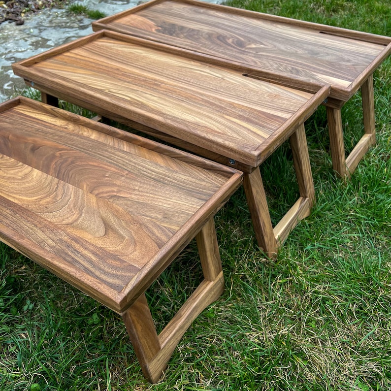 May include: Three wooden folding lap desks with a rich brown finish. Each desk has a rectangular top with a raised edge and angled legs. The desks are nested together, showcasing the varying sizes and wood grain patterns.
