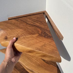 May include: A set of wooden corner shelves with a natural wood grain finish. The shelves are triangular and appear to be mounted on a white wall. The wood has a warm, reddish-brown hue. The shelves are held by a wooden support.
