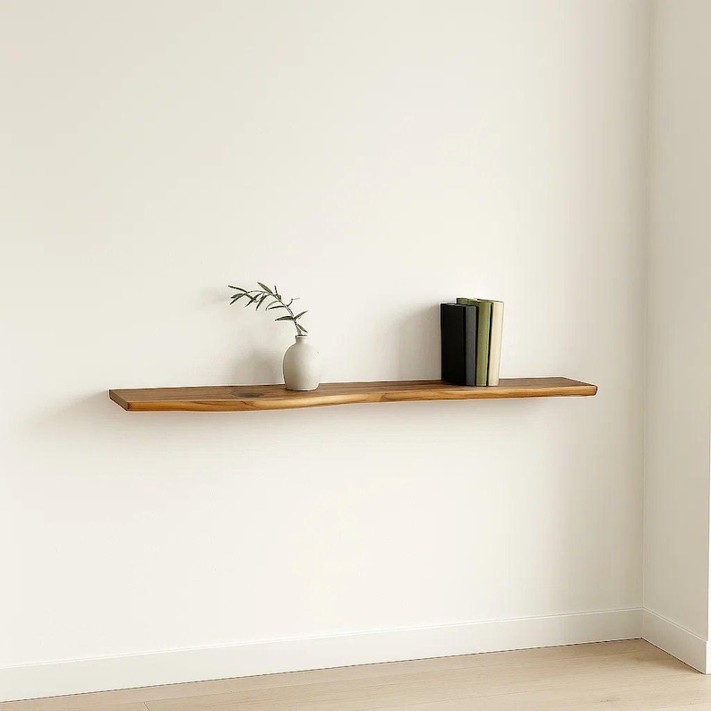 May include: A floating wooden shelf with a natural edge, holding a small white vase with greenery and a stack of books. The shelf is mounted on a white wall, creating a minimalist aesthetic. The shelf is approximately 48 inches long.