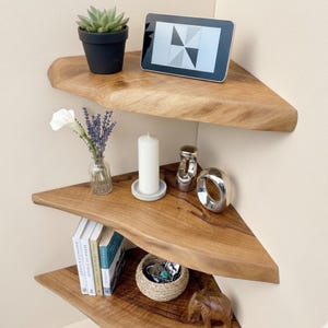 Puede incluir: Tres estanterías de esquina de madera con bordes naturales, que muestran una suculenta en una maceta negra, una tableta, una vela, libros y objetos decorativos. Las estanterías son de madera de color marrón claro.