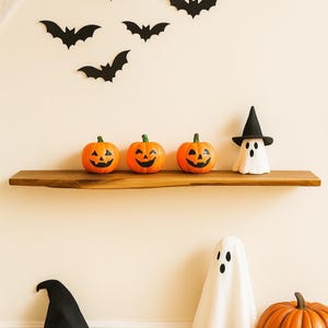 May include: A wooden shelf displays three orange jack-o'-lanterns with carved faces and a white ghost figurine wearing a black witch hat. Black bat cutouts are affixed to the wall above, creating a Halloween theme.