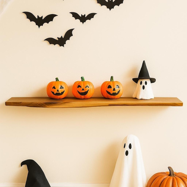 May include: A wooden shelf displays three orange jack-o'-lanterns with carved faces and a white ghost figurine wearing a black witch hat. Black bat cutouts are affixed to the wall above, creating a Halloween theme.