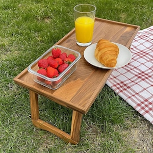 May include: A rectangular wooden breakfast tray with folding legs, featuring a clear glass container filled with fresh red strawberries, a white plate holding a golden-brown croissant, and a tall glass of bright orange juice on the surface.