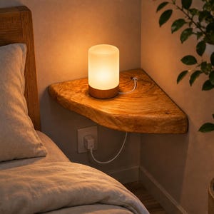 May include: A cylindrical table lamp with a frosted white glass shade and a light wood base, resting on a triangular live-edge wooden corner shelf. The lamp is connected by a white cord to a wall outlet located directly below the shelf.