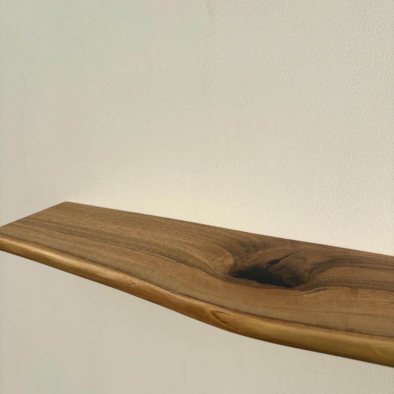 May include: A natural wood floating shelf with a live edge. The shelf is a warm brown color with darker grain patterns and a knot detail. The shelf is approximately 30 inches long and has a smooth, finished surface.