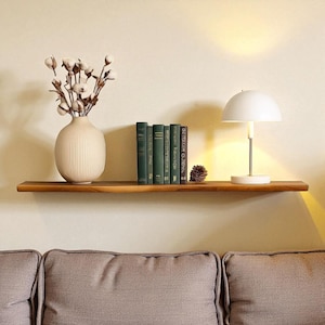 May include: A wooden floating shelf displays a cream-colored vase with cotton stems, several green books, a white lamp, and a pinecone. The shelf is mounted on a beige wall above a gray sofa, creating a cozy, decorative space.