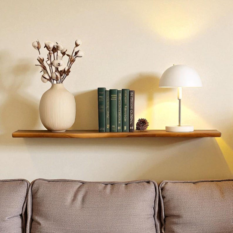May include: A wooden floating shelf displays a cream-colored vase with cotton stems, several green books, a white lamp, and a pinecone. The shelf is mounted on a beige wall above a gray sofa, creating a cozy, decorative space.