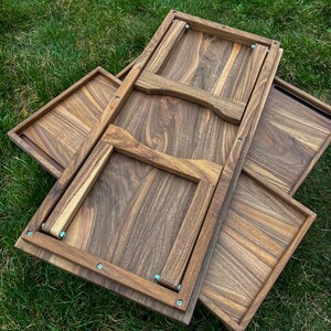 May include: Wooden serving tray set with a folding table. The trays and table are made of a dark brown wood with visible grain. The table is partially folded, revealing its frame and hinges. The set is placed on green grass.