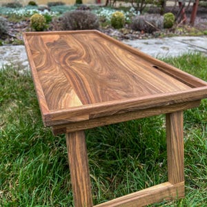 May include: A wooden table with a natural wood finish. The table has a rectangular top and four legs. The table is sitting on a bed of green grass.