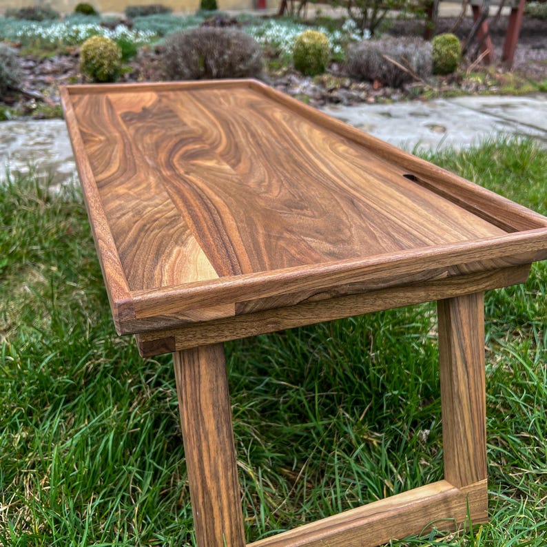 May include: A wooden table with a natural wood finish. The table has a rectangular top and four legs. The table is sitting on a bed of green grass.