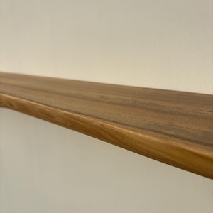 May include: A solid wood floating shelf with a natural finish. The shelf is a warm brown color and has a smooth, polished surface. The shelf is mounted on a white wall.