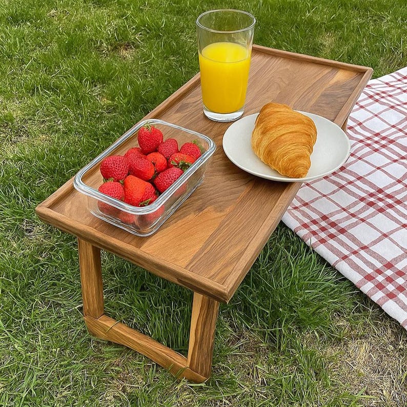 May include: A wooden picnic table with a glass of orange juice, a plate with a croissant, and a glass container of strawberries. The table is set on a grassy area with a red and white checkered blanket.