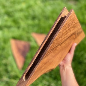 May include: A wooden cutting board with a handle is held up, showcasing its natural wood grain. The board has a slot cut into it. Two additional wooden triangles are blurred in the background, set against a backdrop of green grass.