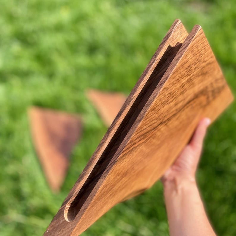 May include: A wooden cutting board with a handle is held up, showcasing its natural wood grain. The board has a slot cut into it. Two additional wooden triangles are blurred in the background, set against a backdrop of green grass.