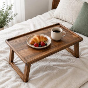 May include: A rectangular wooden breakfast tray with foldable legs featuring a dark brown wood grain finish. On the tray sits a white ceramic plate holding a golden-brown croissant, two red strawberries, and a blueberry, alongside a white mug.
