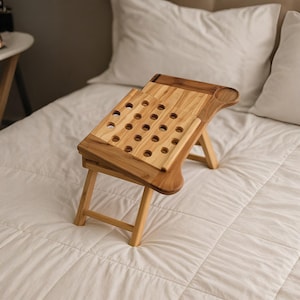 May include: A foldable bamboo laptop desk with an adjustable tilting surface featuring circular ventilation cutouts. The desk includes a flat side section with a circular indentation for a cup and four sturdy legs for stable placement on a bed.