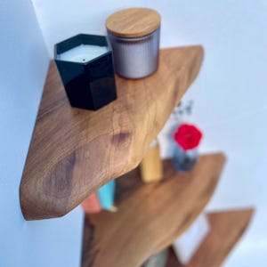 May include: A set of three wooden floating shelves with a natural edge. The top shelf holds a black geometric candle holder with a white candle and a gray glass jar with a wooden lid. A small vase with a red flower is on the bottom shelf.