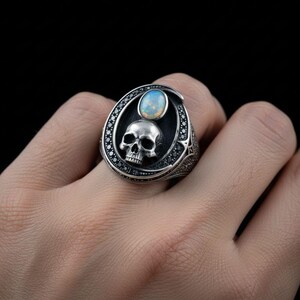 Op de afbeelding: Een zilveren ring met een centrale ovale opaal edelsteen boven een gedetailleerd schedelmotief. De ringkop is omlijst door een rand van kleine zwarte steentjes die in het metaal zijn gezet, met een geoxideerde afwerking voor extra textuur.