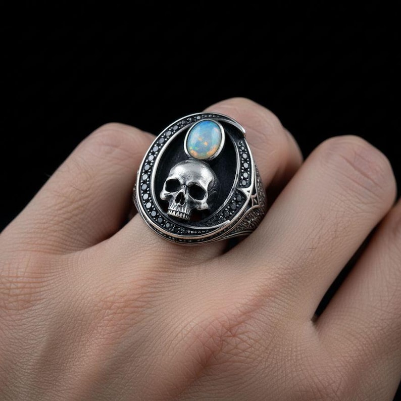 Op de afbeelding: Een zilveren ring met een centrale ovale opaal edelsteen boven een gedetailleerd schedelmotief. De ringkop is omlijst door een rand van kleine zwarte steentjes die in het metaal zijn gezet, met een geoxideerde afwerking voor extra textuur.