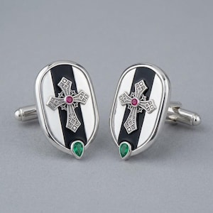 May include: A pair of oval silver cufflinks featuring a central black vertical stripe. Each cufflink displays an ornate silver cross with a small round red gemstone at its centre and a single pear-shaped green gemstone at the bottom edge.