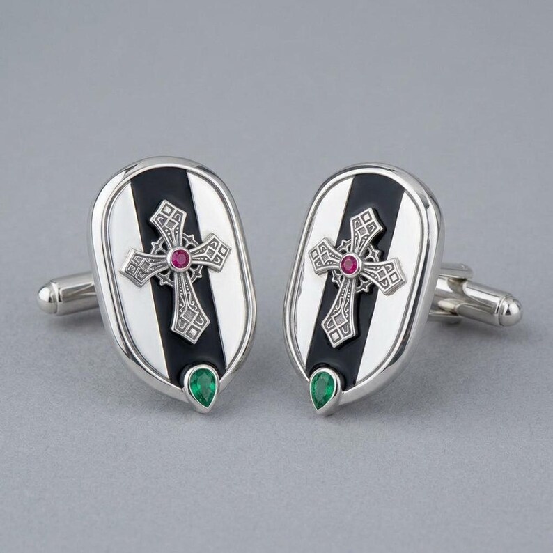 May include: A pair of oval silver cufflinks featuring a central black vertical stripe. Each cufflink displays an ornate silver cross with a small round red gemstone at its centre and a single pear-shaped green gemstone at the bottom edge.