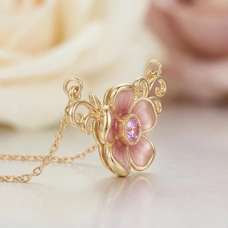 May include: A gold-colored necklace featuring a pink flower pendant with a central pink gemstone. The flower has delicate pink petals and ornate gold detailing. The chain is fine and gold, creating an elegant jewelry piece.