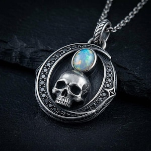 Peut inclure: Un pendentif en argent avec un cadre circulaire incrust&eacute; de petites pierres noires. &Agrave; l'int&eacute;rieur du cadre se trouve un cr&acirc;ne en argent d&eacute;taill&eacute; surmont&eacute; d'un cabochon ovale en opale iris&eacute;e. Le pendentif est suspendu &agrave; une cha&icirc;ne en argent.