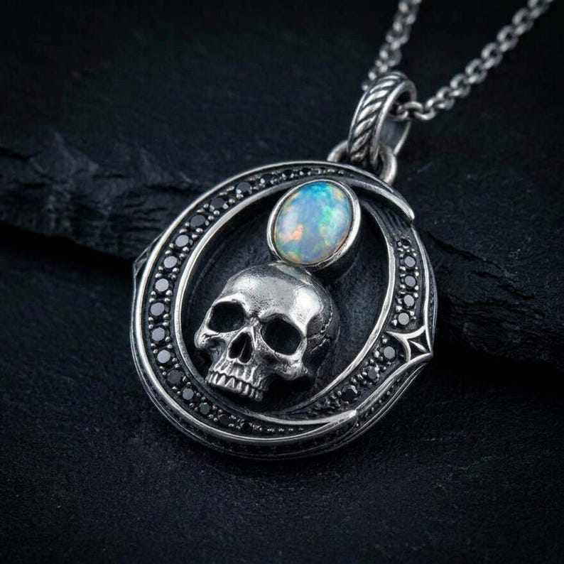 Peut inclure: Un pendentif en argent avec un cadre circulaire incrust&eacute; de petites pierres noires. &Agrave; l'int&eacute;rieur du cadre se trouve un cr&acirc;ne en argent d&eacute;taill&eacute; surmont&eacute; d'un cabochon ovale en opale iris&eacute;e. Le pendentif est suspendu &agrave; une cha&icirc;ne en argent.