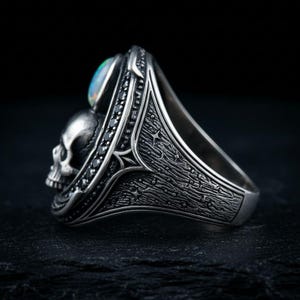 Op de afbeelding: Een zilveren ring met een gedetailleerd schedelmotief en een ovale iriserende opaal cabochon. De band is versierd met kleine zwarte steentjes en ingewikkelde ge&euml;tste patronen die doen denken aan een sterrenhemel, met een gepolijste afwerking.