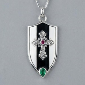 May include: A silver shield-shaped pendant necklace featuring a central black vertical stripe. An ornate silver cross with a small round red gemstone at its centre is layered over the black stripe, with a pear-shaped green gemstone at the base.