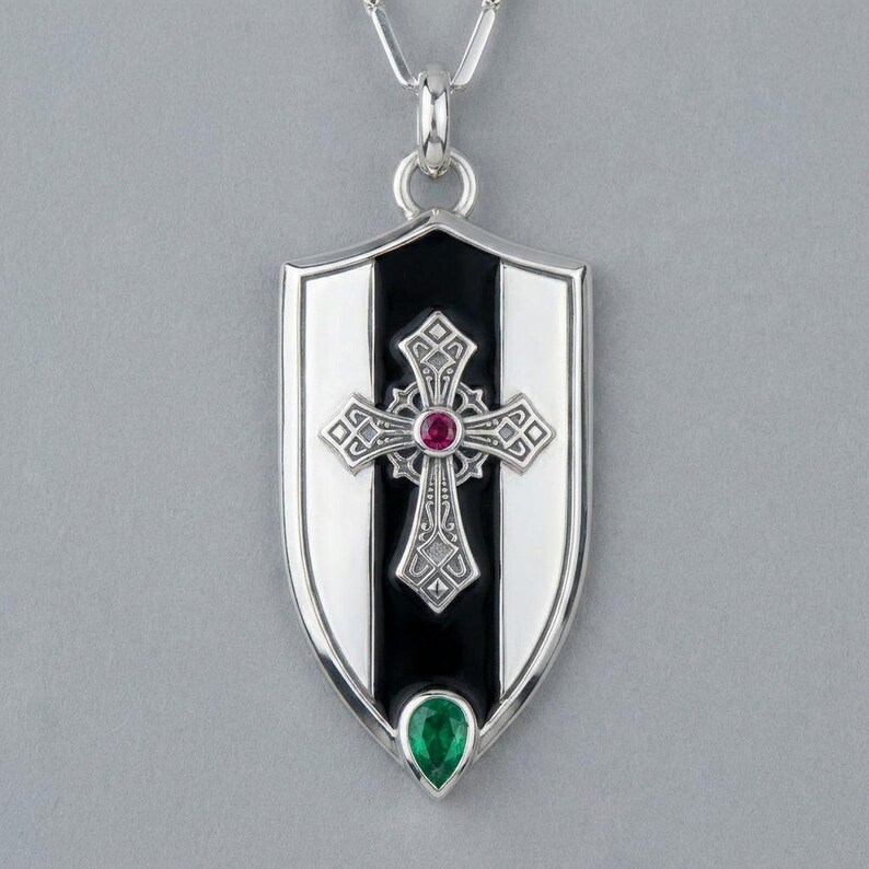 May include: A silver shield-shaped pendant necklace featuring a central black vertical stripe. An ornate silver cross with a small round red gemstone at its centre is layered over the black stripe, with a pear-shaped green gemstone at the base.