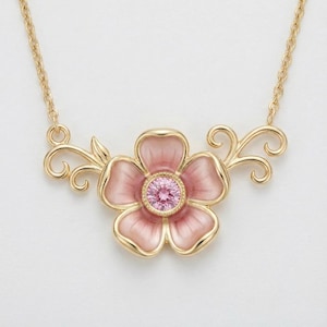 May include: A gold-toned necklace featuring a pink flower pendant with a central pink gemstone. The flower has five petals with a gold outline. The necklace chain and decorative flourishes on either side of the flower are also gold.
