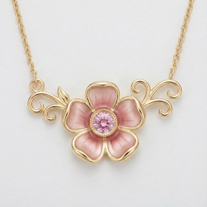 May include: A gold-toned necklace featuring a pink flower pendant with a central pink gemstone. The flower has five petals with a gold outline. The necklace chain and decorative flourishes on either side of the flower are also gold.