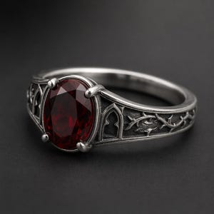 May include: A silver-toned ring featuring a large, oval-cut deep red gemstone held by four prongs. The band is detailed with embossed gothic-style archways and intricate thorny vine patterns, creating a textured, metallic appearance throughout the piece.