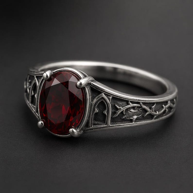 May include: A silver-toned ring featuring a large, oval-cut deep red gemstone held by four prongs. The band is detailed with embossed gothic-style archways and intricate thorny vine patterns, creating a textured, metallic appearance throughout the piece.