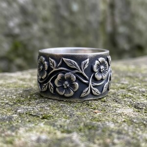 Pu&ograve; includere: Un anello largo color argento con un design floreale. L'anello presenta fiori e foglie in rilievo attorno alla fascia. L'anello &egrave; posto su uno sfondo naturale e strutturato.