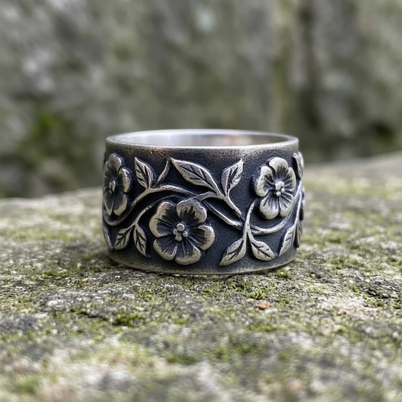 Pu&ograve; includere: Un anello largo color argento con un design floreale. L'anello presenta fiori e foglie in rilievo attorno alla fascia. L'anello &egrave; posto su uno sfondo naturale e strutturato.