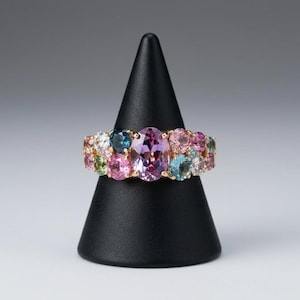 Puede incluir: Un anillo de oro con una gran piedra preciosa central de corte ovalado en color p&uacute;rpura, rodeada por un grupo de piedras m&aacute;s peque&ntilde;as de corte redondo y multicolor, incluyendo gemas rosas, azules, verdes y transparentes en una banda de oro.