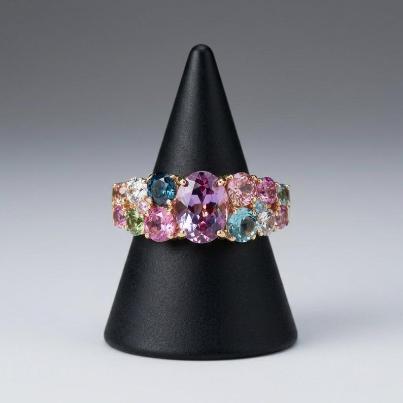 Puede incluir: Un anillo de oro con una gran piedra preciosa central de corte ovalado en color p&uacute;rpura, rodeada por un grupo de piedras m&aacute;s peque&ntilde;as de corte redondo y multicolor, incluyendo gemas rosas, azules, verdes y transparentes en una banda de oro.