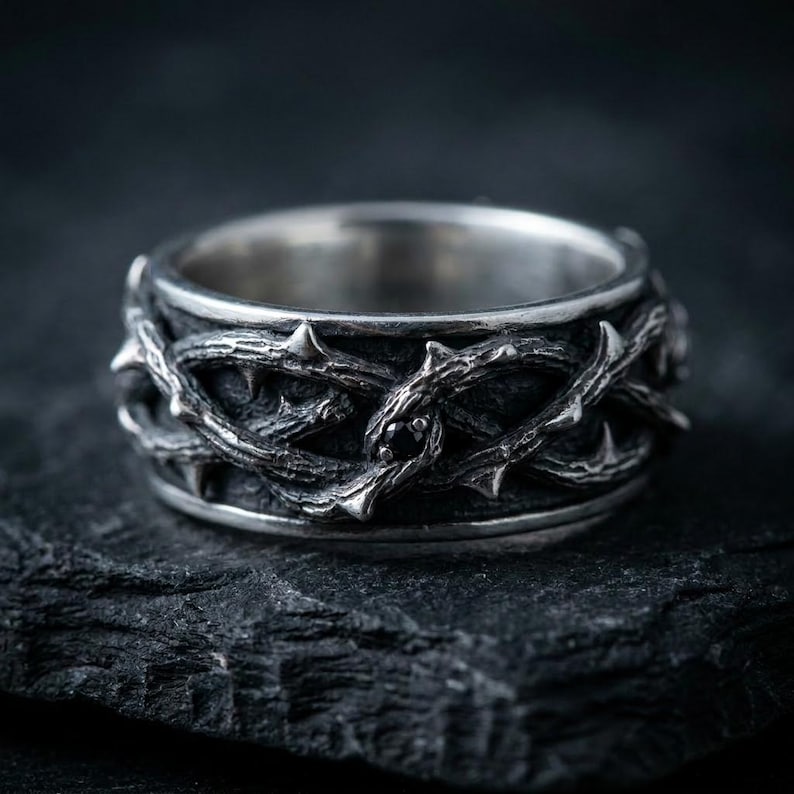 May include: A silver ring featuring a textured, three-dimensional band designed to resemble intertwined thorny branches. The metal has a dark, oxidized finish in the recessed areas, and a single small, round black gemstone is set within the design.