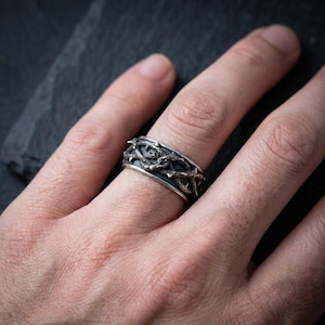 May include: A silver-toned metal ring featuring a continuous, raised relief design of intertwined thorny branches. The band has a dark, oxidized finish within the recessed areas to highlight the intricate texture of the thorns around the circumference.