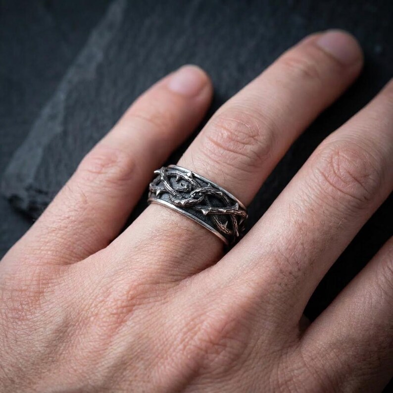 May include: A silver-toned metal ring featuring a continuous, raised relief design of intertwined thorny branches. The band has a dark, oxidized finish within the recessed areas to highlight the intricate texture of the thorns around the circumference.