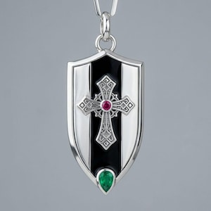 May include: A silver shield-shaped pendant featuring a central black vertical stripe, an ornate silver cross with a round red gemstone at its centre, and a pear-shaped green gemstone set at the bottom point of the shield, suspended from a silver bail.