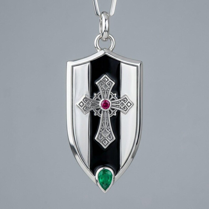May include: A silver shield-shaped pendant featuring a central black vertical stripe, an ornate silver cross with a round red gemstone at its centre, and a pear-shaped green gemstone set at the bottom point of the shield, suspended from a silver bail.