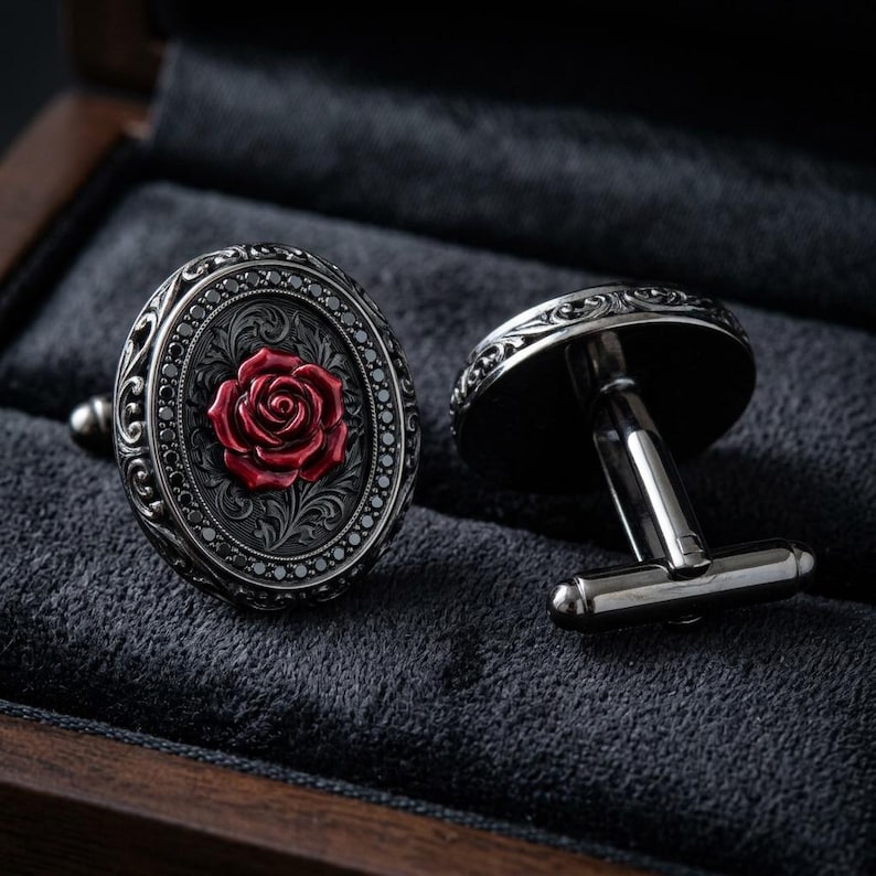 May include: A pair of oval cufflinks featuring a dark metallic finish with intricate scrollwork engravings surrounding a central, raised red rose emblem. The cufflinks have a polished, reflective surface and a standard T-bar toggle mechanism.