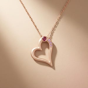 May include: A rose gold heart pendant necklace. The open heart design features a small, round, red gemstone at the top and a cluster of small pink gemstones along one side. The chain is delicate and matches the pendant's colour.