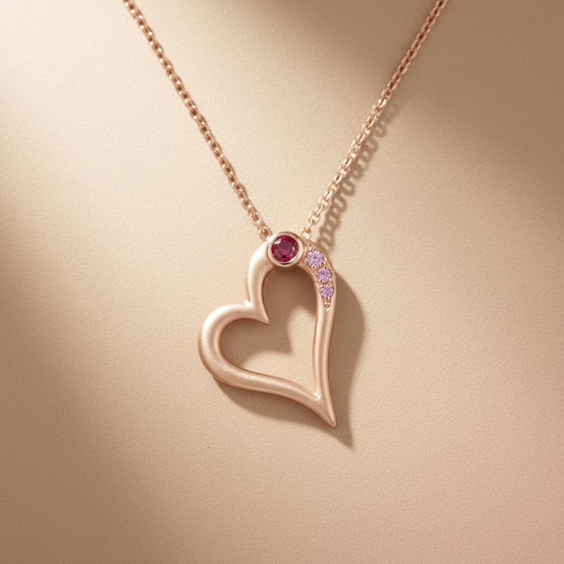 May include: A rose gold heart pendant necklace. The open heart design features a small, round, red gemstone at the top and a cluster of small pink gemstones along one side. The chain is delicate and matches the pendant's colour.