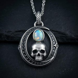 Peut inclure: Un collier pendentif en argent repr&eacute;sentant un cr&acirc;ne humain central surmont&eacute; d'un cabochon d'opale ronde et iris&eacute;e. Le cr&acirc;ne est encadr&eacute; par une forme de croissant de lune incrust&eacute;e de petites pierres noires, suspendu &agrave; une cha&icirc;ne en argent textur&eacute;e.