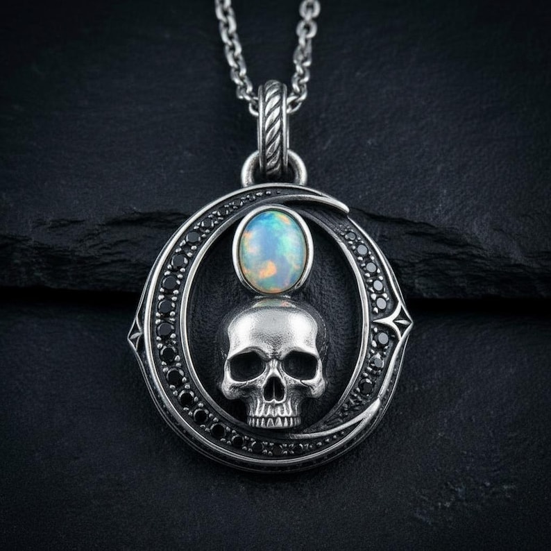 Peut inclure: Un collier pendentif en argent repr&eacute;sentant un cr&acirc;ne humain central surmont&eacute; d'un cabochon d'opale ronde et iris&eacute;e. Le cr&acirc;ne est encadr&eacute; par une forme de croissant de lune incrust&eacute;e de petites pierres noires, suspendu &agrave; une cha&icirc;ne en argent textur&eacute;e.