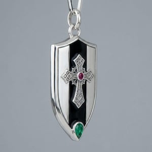 May include: A silver pendant shaped like a shield, featuring a central black vertical stripe with an ornate silver cross overlay. The cross is accented with a small round red gemstone at its centre, and a teardrop-shaped green gemstone sits below.